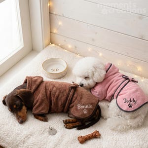 Könnte beinhalten: Zwei Hunde mit Kapuzenbademänteln liegen auf einer flauschigen, weißen Oberfläche. Einer trägt einen braunen Bademantel mit der Aufschrift "Bonnie", der andere einen rosa Bademantel mit "Teddy". Eine Keramikschale mit "SIP" ist ebenfalls zu sehen.
