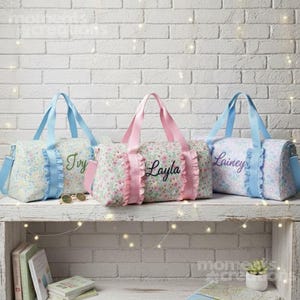 May include: Three floral print duffel bags with personalized names embroidered in cursive. The bags have ruffled trim and matching colored handles. One bag has a blue floral print with blue handles and the name "Ivy". Another has a pink floral print with pink handles and the name "Layla". The third bag has a blue floral print with blue handles and the name "Lainey".