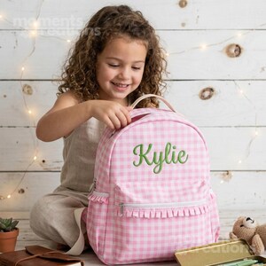 Pode incluir: Mochila rosa xadrez com o nome "Kylie" bordado em verde. A mochila tem uma alça superior, um bolso frontal com zíper e uma borda com babados. Uma criança está segurando a mochila.