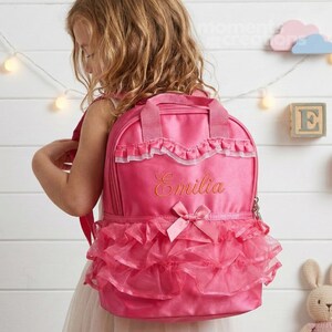 May include: A pink backpack with a tutu-style skirt and the name "Emilia" embroidered in gold. The backpack has a top handle, a bow, and white lace trim. The bag is designed for a child.