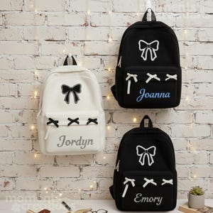Peut inclure: Trois sacs à dos accrochés à un mur de briques. L'un est blanc avec un nœud noir et le nom "Jordyn" en gris. Les deux autres sont noirs, avec des nœuds blancs et les noms "Joanna" et "Emory" en bleu et gris. Chacun a une poche avant.