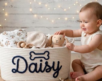 Könnte beinhalten: Ein weißer Korb aus geflochtenem Seil mit dem Namen Daisy in dunkelblauer Schreibschrift und einem Knochen-Symbol darüber. Der Korb enthält ein Tuch mit Blumenmuster, eine beige Strickhaube und ein beiges Stoffhasen-Spielzeug.