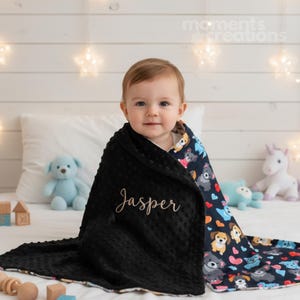 May include: A baby wrapped in a black blanket embroidered with the name "Jasper". The blanket has a textured surface and a patterned edge featuring dogs and hearts. A blue teddy bear and a unicorn toy are visible in the background.