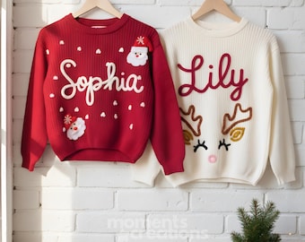 May include: Two long-sleeve knit sweaters hanging on wooden hangers. The red sweater features the name Sophia in white script, two Santa Claus faces, and white dots. The cream sweater features the name Lily in red script and a reindeer face.
