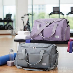 May include: Two duffel bags, one lavender and one gray, sit on a bench. Both bags have multiple pockets, shoulder straps, and the text "Olivia" on the gray bag. A blue yoga mat, water bottle, and exercise bands are nearby.