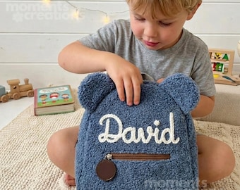 May include: A blue teddy bear-shaped backpack made of textured sherpa fleece fabric. The front features the name David embroidered in white script lettering above a brown zippered pocket with a round brown zipper pull.
