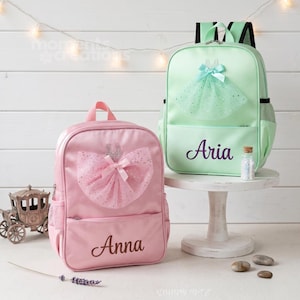 May include: Two children's backpacks, one pink and one mint green, are displayed. The pink backpack features a tutu design and the name "Anna". The green backpack has a tutu design and the name "Aria". Both have side pockets.