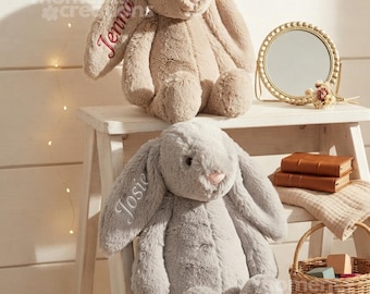 May include: Two plush stuffed bunnies with long floppy ears. The top bunny is beige with the name Jenna embroidered in red on its ear. The bottom bunny is gray with the name Josie embroidered in white on its ear. Both have soft fur.