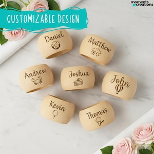 Può includere: Un set di anelli portatovaglioli in legno personalizzati, ognuno inciso con un nome e una piccola illustrazione. I nomi includono "Daniel" e "Matthew", con icone come una ciambella e nuvole di pioggia. È visibile anche il testo "CUSTOMIZABLE DESIGN".