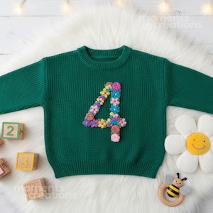 May include: A green knitted jumper featuring a floral-decorated number "4" on the front. The image includes wooden number blocks, a daisy cushion, and a wooden bee-shaped rattle. The jumper is a children's garment.