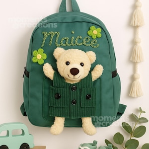 May include: Green backpack with a teddy bear pocket and the name "Maicee" embroidered in green. The bear wears a green vest. The backpack has green flower accents.