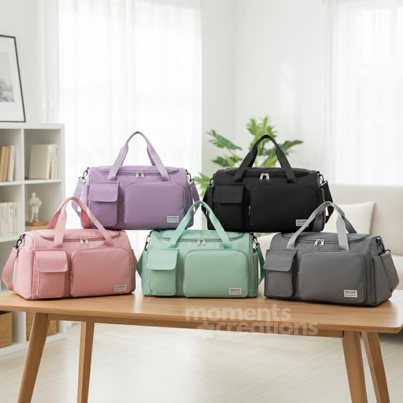 May include: Five travel bags in various colors: lavender, pink, mint green, black, and gray. Each bag has a rectangular shape, two front pockets, top handles, and a shoulder strap. The bags are displayed on a wooden table.