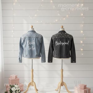K&ouml;nnte beinhalten: Zwei Jeansjacken auf einer Pr&auml;sentation. Eine ist hellblau mit dem Text "Bride to be" und Perlenverzierungen. Die andere ist dunkelgrau mit dem Text "Bridesmaid" und &auml;hnlichen Verzierungen. Beide stehen auf Holzst&auml;ndern.