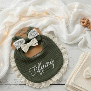 May include: An olive green baby bib with the name "Tiffany" embroidered in white script. The bib has a cream-colored lace trim and is paired with a matching headband and bow. A wooden toy and a stack of cream-colored cloths are also visible.