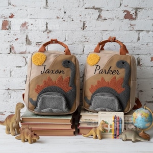 以下が含まれることがあります： 「Jaxon」と「Parker」の名前が刺繍された、ベージュと茶色の恐竜をテーマにした2つのバックパック。各バックパックは、オレンジ色のアクセントと黄色の太陽が付いた灰色の恐竜のデザインが特徴です。バックパックは本の上に置かれ、おもちゃの恐竜と地球儀が近くにあります。