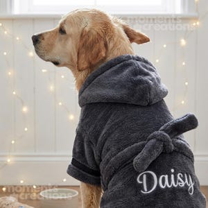 Może przedstawiać: Golden retriever w szarym szlafroku z kapturem i pasującym paskiem. Szlafrok ma czarne wykończenia i imię "Daisy" wyhaftowane na biało. Pies jest w pomieszczeniu, z miękkim oświetleniem.