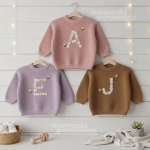 May include: Three children's jumpers in pink, lavender, and brown, each with a floral initial and bee accents. The jumpers are hanging on wooden hangers against a white wood-panelled wall. The brand name "moments creations" is visible.