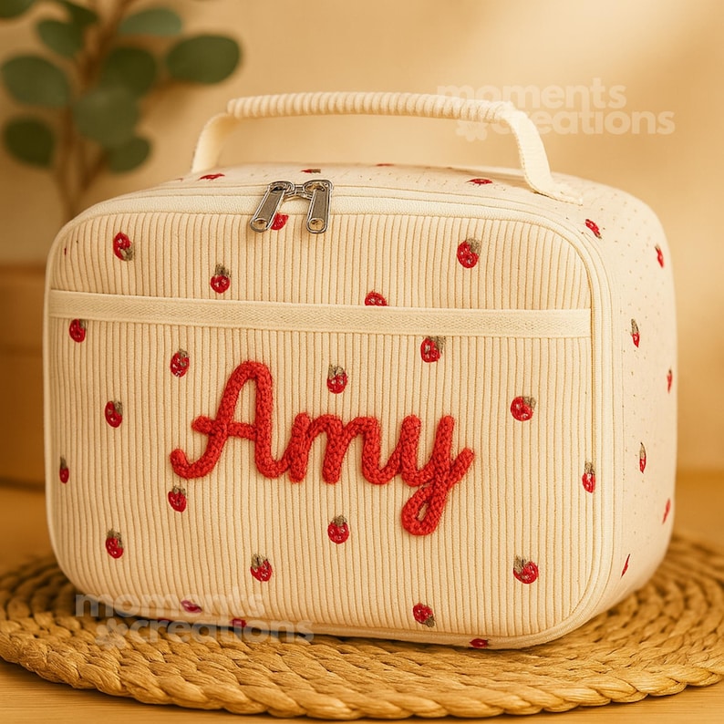 May include: Cream-colored lunch bag with a handle and zipper closure. The bag is decorated with red embroidered strawberries and the name "Amy" in red script. The bag has a front pocket and is sitting on a woven mat.
