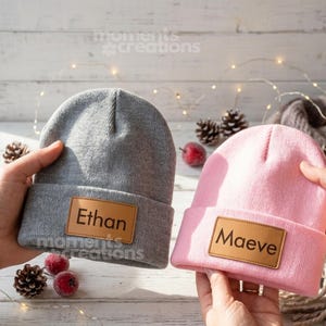 Puede incluir: Un gorro de punto gris con un parche de cuero marrón grabado con el nombre Ethan y un gorro de punto rosa con un parche de cuero marrón grabado con el nombre Maeve. Ambos gorros cuentan con un borde doblado y tejido texturizado.