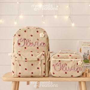 May include: A beige backpack and lunch bag set with a strawberry pattern. The name "Olivia" is embroidered in pink on both items. Each bag has a white flower accent.