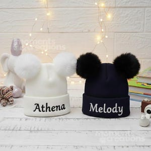 Op de afbeelding: Twee gebreide mutsen met dubbele pompons. De witte muts heeft de naam Athena in het zwart geborduurd, terwijl de donkerblauwe muts de naam Melody in het wit geborduurd heeft. Beide mutsen hebben een omgeslagen boord.