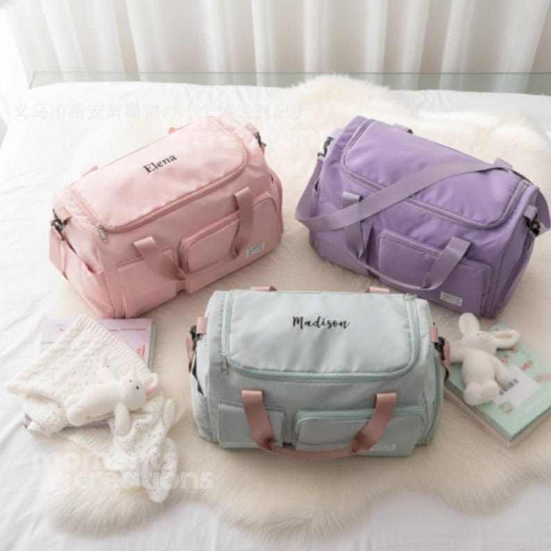 May include: Three personalized baby bags in pastel colors: pink, lavender, and mint green. Each bag has a name printed on it: "Elena" on the pink bag, "Madison" on the mint green bag. The bags have multiple pockets and handles.