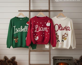 May include: Three knit sweaters hanging on a wooden ladder. The green sweater features the name Isabell and a reindeer. The red sweater features the name Liam and Santa Claus. The white sweater features the name Olivia and a reindeer.