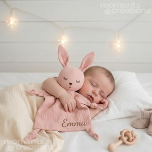 Könnte beinhalten: Ein schlafendes Baby kuschelt mit einem rosa Hasen-Schmusetuch, auf dem der Name "Emma" gestickt ist. Der Hase hat lange Ohren und geschlossene Augen. Ein Holzbeißring und Babyschuhe sind ebenfalls zu sehen.