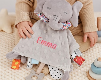 May include: A gray plush elephant security blanket with a dimpled texture, featuring the name Emma embroidered in pink thread. The blanket is adorned with a colorful floral collar and various patterned fabric tags, including a star-shaped teether.