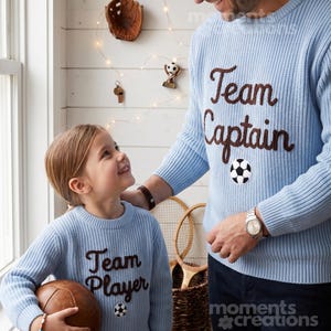 Peut inclure: Deux pulls en tricot bleu clair, l'un avec "Team Captain" et un ballon de football, l'autre avec "Team Player" et un ballon de football. Les pulls sont portés par une personne et un enfant, respectivement.
