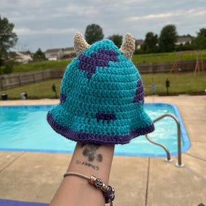 May include: A blue and purple crocheted hat with two beige horns. The hat is shaped like a monster with a wide brim.