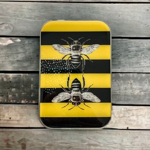 May include: A silver metal rectangle with a black and yellow striped design. Two black and white bees are depicted on the rectangle. The bees are facing forward with their wings spread out.