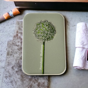 May include: A rectangular, sage green coaster with a dandelion-like flower design. The flower is a sphere of tiny white blossoms atop a green stem. The words "Random Buttons" are printed vertically on the left side.