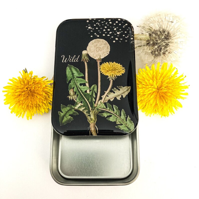 May include: A black metal tin with a dandelion illustration and the word "Wild". The tin is open, revealing a silver interior. Bright yellow dandelions and a white dandelion puff are arranged around the tin.