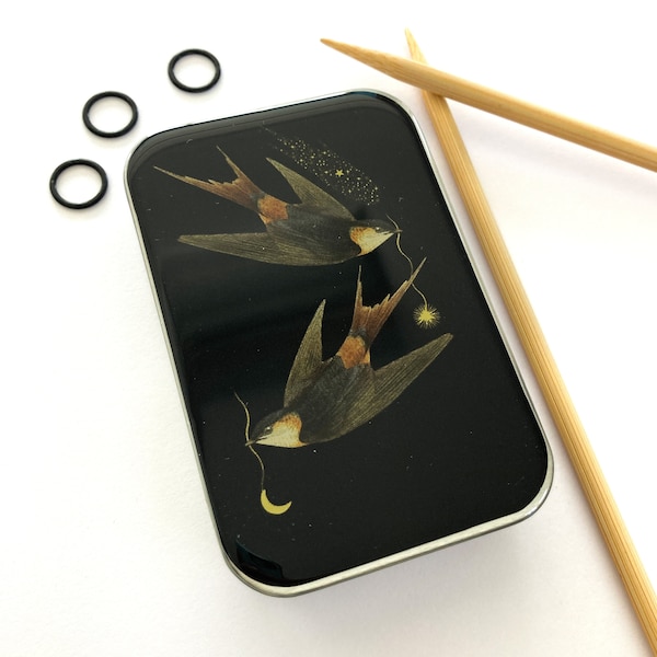 May include: A black metal box with a silver trim, featuring a design of two swallows flying against a black background with gold stars and a crescent moon.