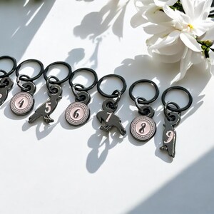 May include: A collection of black metal stitch markers with charms. Each charm features a number from 4 to 9, and a bird or circular design with a pink border. The stitch markers are on a white surface.