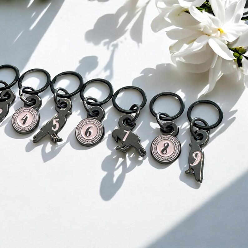 May include: A collection of black metal stitch markers with charms. Each charm features a number from 4 to 9, and a bird or circular design with a pink border. The stitch markers are on a white surface.