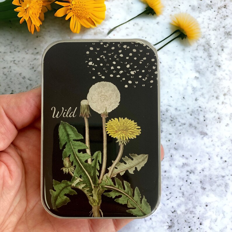 May include: A rectangular black tin with a dandelion design. The tin features a yellow dandelion flower, a white dandelion seed head, and the word "Wild" in white script. The background is black with white speckles.