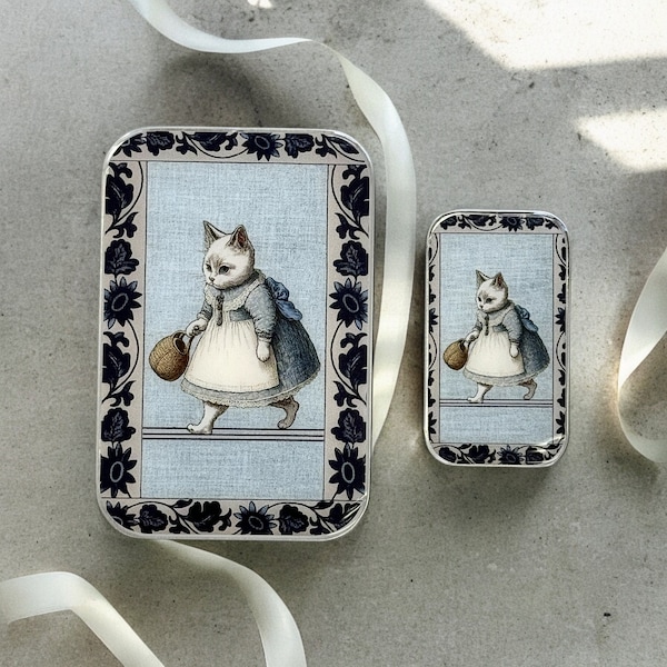 Op de afbeelding: Twee vintage blikken doosjes met een blauw en wit bloemenontwerp. Elk doosje heeft een zwart-witte illustratie van een kat die een emmer draagt.