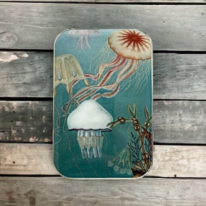 Może przedstawiać: Podkładka pod kubek ze szkła w kolorze niebieskim i zielonym z vintage ilustracją meduz. Meduzy są różowe, białe i pomarańczowe. Podkładka ma białą ramkę.