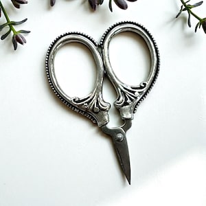 May include: Silver embroidery scissors with decorative handles and a detailed edge. The scissors are open, showing the sharp blades. The design is reminiscent of vintage sewing tools, ideal for crafting and needlework projects.