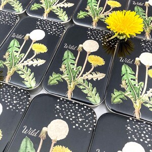 May include: Rectangular black tins with a dandelion design. The tins feature a watercolor illustration of dandelions with yellow and white seed heads, green leaves, and the word "Wild". A fresh yellow dandelion is placed on top.