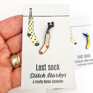Puede incluir: Un marcador de punto de metal negro con forma de calcetín con un corazón rojo en la punta. El marcador está unido a un clip de metal negro. La tarjeta dice "Lost Sock Stitch Marker A Firefly Notes Exclusive".