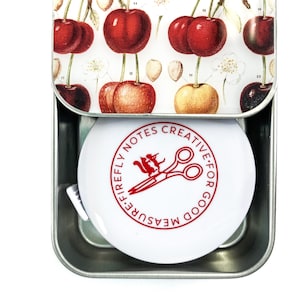 May include: A white metal tin with a red and white label that says "Firefly Notes Creative: For Good Measure". The label has a red illustration of a squirrel holding a pair of scissors. The tin is open and a white tape measure is inside.