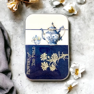 May include: A blue and white porcelain teapot with floral designs sits on a blue background with gold accents. The text "Flowers of the Field" is printed on the side of the tin.