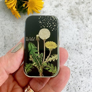 K&ouml;nnte beinhalten: Eine silberne Metalldose mit schwarzem Hintergrund und einer Grafik von drei L&ouml;wenzahnpflanzen. Der Text "Wild" ist in Wei&szlig; auf die Dose gedruckt.