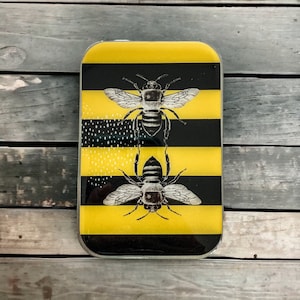 May include: A silver metal rectangle with a black and yellow striped design. Two black and white bees are depicted on the rectangle. The bees are facing forward with their wings spread out.