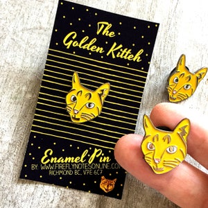 May include: Two yellow enamel pins featuring a cartoon cat with the text "The Golden Kitteh" and "Enamel Pin" on a black and yellow striped background. The text below the cat reads "By: www.fireflynotesonline.com Richmond BC, V7E 6C7"