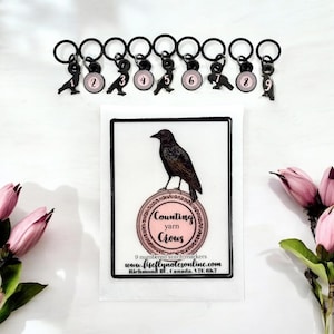 以下が含まれることがあります： 黒い金属製のステッチマーカー9個セット。カラスのチャームと番号付きのピンク色の円が付いています。画像には、カラスのイラストと「Counting Yarn Crows」というテキストが書かれたカードも含まれています。