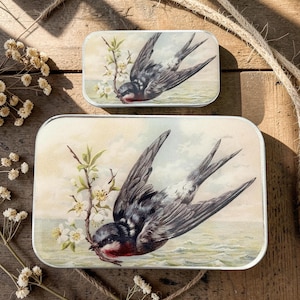 Peut inclure: Deux boîtes métalliques rectangulaires avec une illustration vintage d'oiseau. La plus grande boîte montre une hirondelle aux ailes déployées, tenant une branche fleurie. La plus petite boîte a le même motif. Le fond est un paysage doux de style aquarelle.